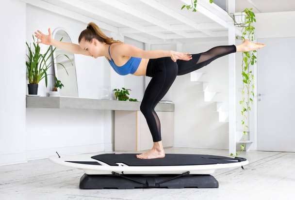 Young woman doing a surfing yoga runner pose balancing on one leg with arms stretched in a high key gym with green plants in a health and fitness concept - Photo, Image