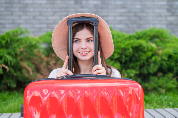 Young beautiful caucasian woman in a hat looks out of a red suitcase with a handle. Happy girl goes on a trip with luggage. Summer vacation concept. - Foto, Bild
