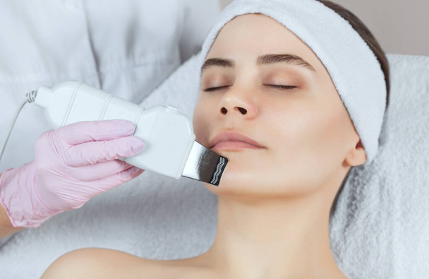 The doctor-cosmetologist makes the apparatus a procedure of ultrasound cleaning of the facial skin of a beautiful, young woman in a beauty salon. Cosmetology and professional skin care. - Photo, Image