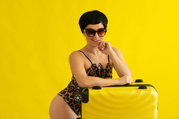 Portrait of an attractive woman in a one-piece leopard swimsuit and sunglasses with a suitcase on a yellow background. Seductive brunette in a monokini is ready to travel to the sea. Summer vacation. - Foto, Imagem