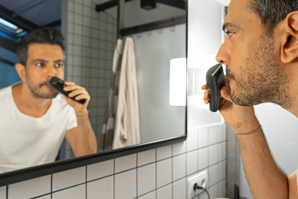 Красивый бородатый мужчина бреет лицо триммером перед зеркалом в ванной - Фото, изображение