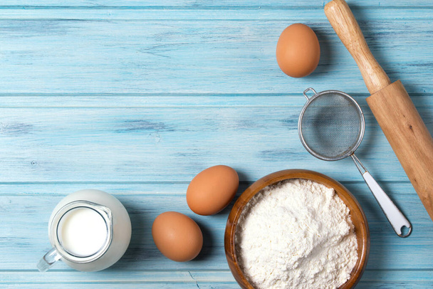 Hozzávalók sütéshez, tejhez, tojáshoz, búzaliszthez és konyhai eszközökhöz kék fa alapon, felülnézetből - Fotó, kép