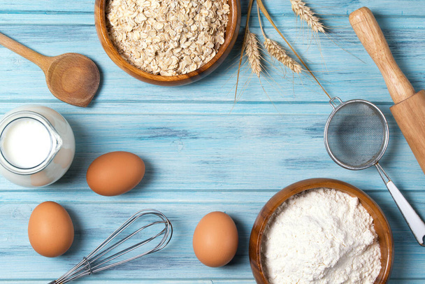 Ingredients for baking, milk, eggs, wheat flour, oats and kitchenware on blue wooden background, top view - Photo, Image