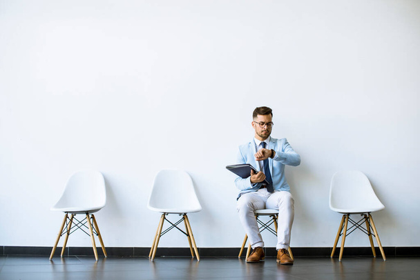 Jeune homme assis à la chaise dans la salle d'attente avec un dossier en main et vérifier le temps avant une entrevue - Photo, image