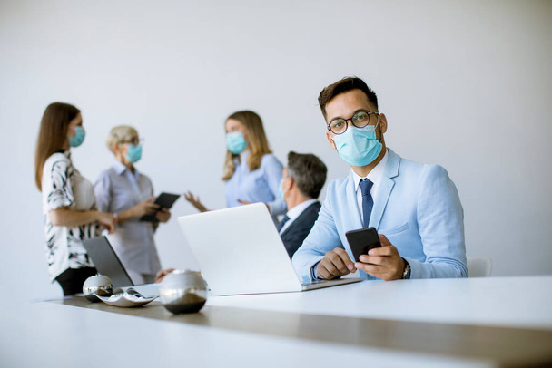 Mladý podnikatel s lékařskou ochrannou maskou pracuje na notebooku a pomocí mobilního telefonu v kanceláři - Fotografie, Obrázek