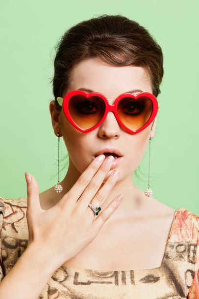 Portrait de jeune femme choquée portant des lunettes de soleil en forme de coeur sur fond vert - Photo, image