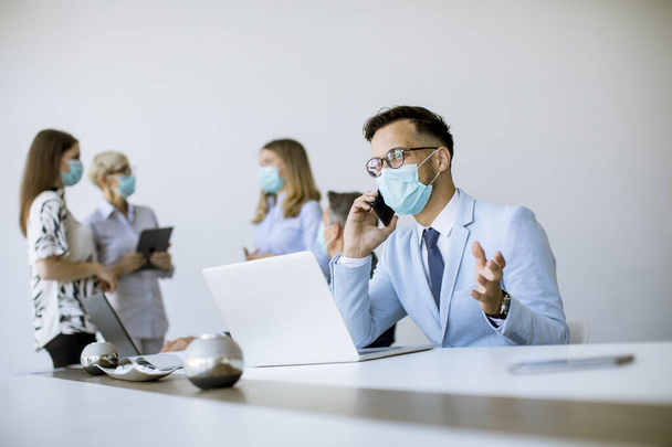 Νεαρός επιχειρηματίας με ιατρική προστατευτική μάσκα εργάζεται σε φορητό υπολογιστή και χρησιμοποιεί κινητό τηλέφωνο στο γραφείο - Φωτογραφία, εικόνα