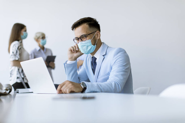 Giovane uomo d'affari con maschera protettiva medica lavora su un computer portatile in ufficio - Foto, immagini