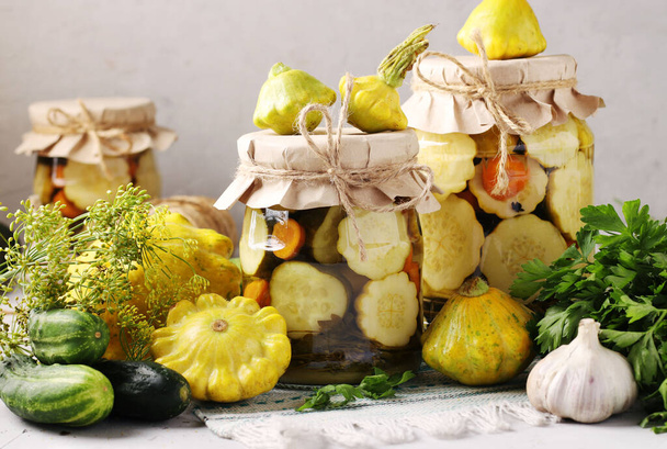 Gepekelde patissons, komkommers met wortelen en knoflook in glazen potjes en verse ingrediënten op een lichtgrijze achtergrond - Foto, afbeelding