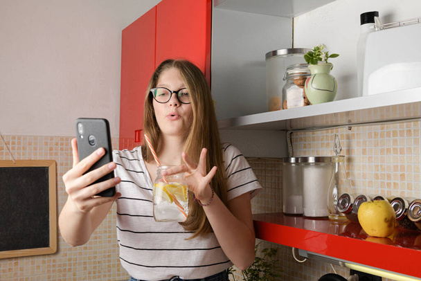 Egy fiatal blogger telefonkamerával beszélget, élő adást tart a közösségi médiában, miközben iszik, miközben a konyhaasztalon ül. Gen z, Milleniumi hobbik. - Fotó, kép