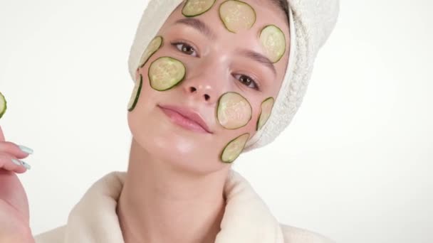 Beautiful tender young girl in a white towel with clean fresh skin posing in front of the camera with a mask of cucumbers on the face. Beauty face. Skin care. - Filmati, video