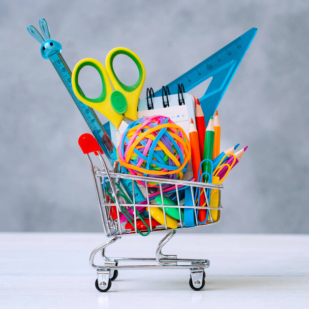 Multi-colored school supplies in a shopping cart on a gray background with copy space for text. - Фото, изображение