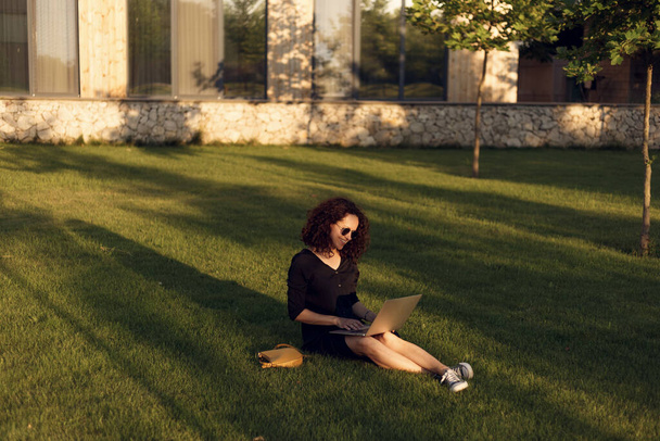 Vrolijke jonge vrouw in een zonnebril zitten op groen gras tijdens de zomer dag met behulp van laptop zittend op gras. - Foto, afbeelding