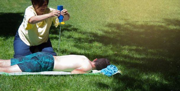 jovem dormindo na grama e banhos de sol e uma mãe pulverizando-o com protetor solar para cuidar de sua pele no verão
 - Foto, Imagem
