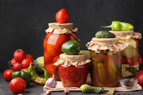 Различные маринованные овощи в стеклянных банках для длительного хранения: перец в томатном соусе, огурцы, помидоры и цуккини темный фон, горизонтальный формат - Фото, изображение
