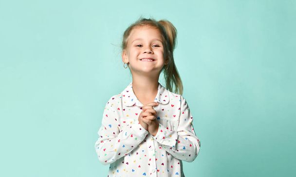 Little blonde schoolgirl with ponytail, in shirt with hearts print. Smiling, posing on blue background. Close up - Fotografie, Obrázek
