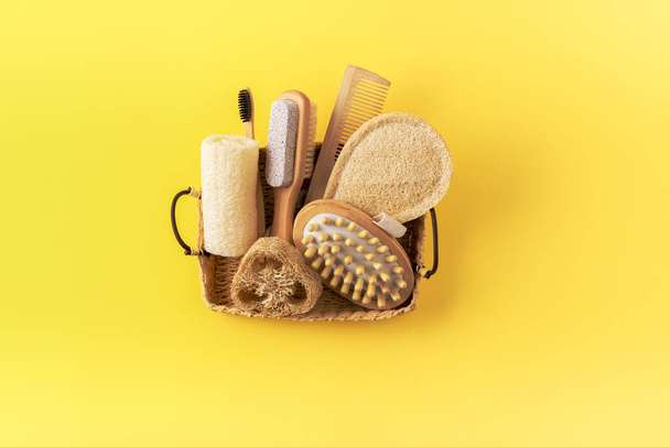 Anti cellulite massager, bamboo toothbrush, loofah sponge, wooden comb, peeling brush in basket on yellow. Eco friendly bathroom accessories. Zero waste concept. Minimalistic style. - Photo, Image