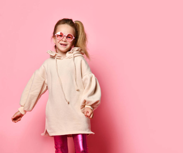 Glamorous little girl in pink glasses posing confidently in a pale pink sweatshirt and glossy leggings. Three quarter length shot, isolated on pink, copy space.  - Фото, зображення