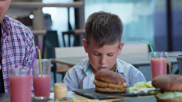 restauration rapide, les enfants masculins affamés avec leur père mangent de savoureux hamburgers de viande juteux avec des frites et du jus de fruits lors d'un dîner en famille au café - Séquence, vidéo