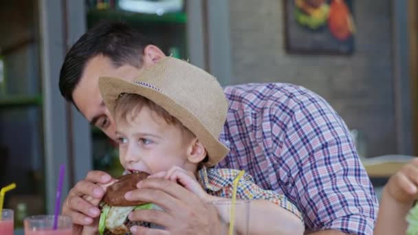 υποσιτισμός, λίγο χαριτωμένο αρσενικό παιδί τρώει fast food χορταστικό μπιφτέκι κρέατος με τον αγαπημένο του πατέρα κατά τη διάρκεια των οικογενειακών διακοπών στο καφέ - Πλάνα, βίντεο