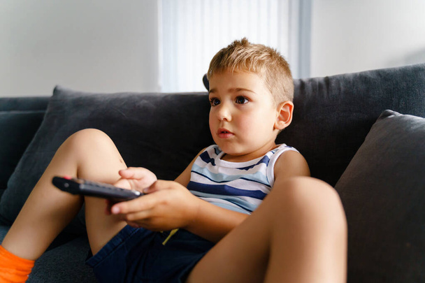 Kisgyerek ül a kanapén otthon kezében TV távirányító - Kisgyermek néz és vált televíziós csatorna - Fiú pihentető otthon nyári nap - gyermekkori szabadidős tevékenység koncepció - Fotó, kép