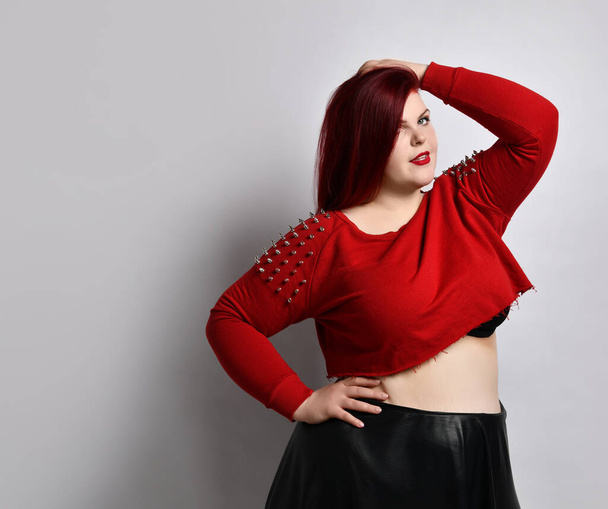 Overweight redhead woman in red spiked top, black leather skirt. Touching face, put hand on hip, posing sideways isolated on white - Foto, immagini
