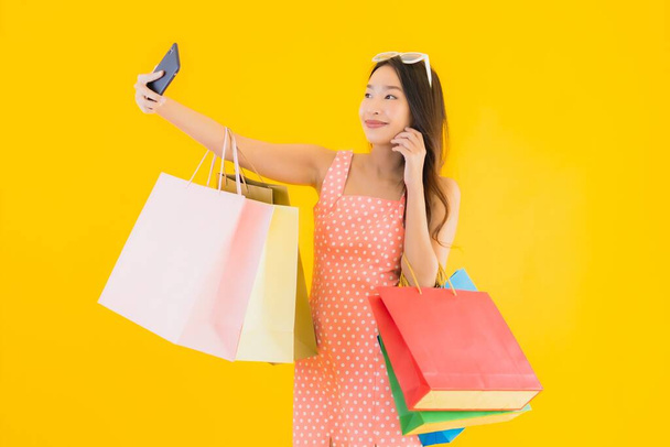 Portrait beautiful young asian woman with colorful shopping bag with smart mobile phone and credit card on yellow isolated background - Photo, Image
