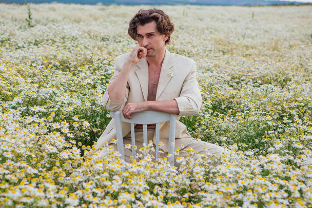 Tall handsome man dressed in a white suit on naked body sitting on a white chair in camomile flowers field - Foto, Imagem