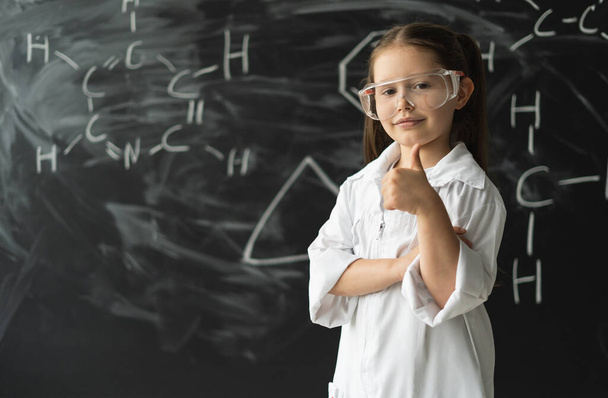Estudante menina fica perto do quadro com fórmulas. Levanta o polegar. Casaco branco e óculos - Foto, Imagem