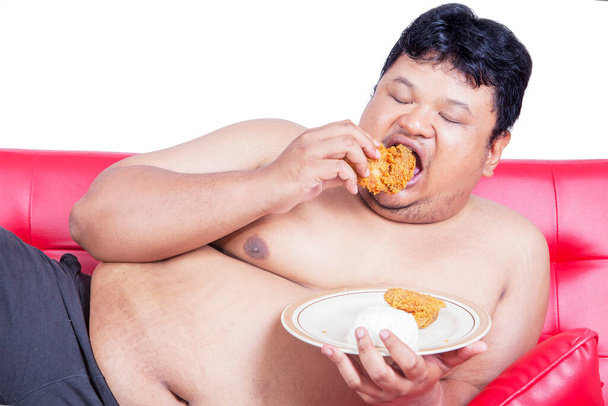 Close up of an obesity man eating a plate of fried chickens while sitting on the couch. Shot in the studio with isolated on white background - Фото, зображення