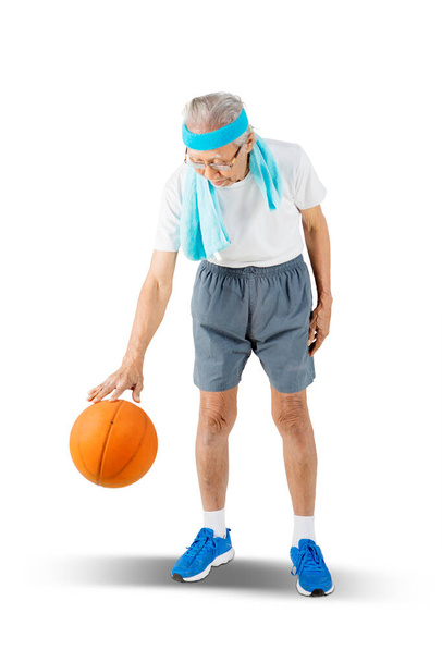 Elderly man wearing sportswear while dribbling a basketball in the studio. Isolated on white background - Photo, image