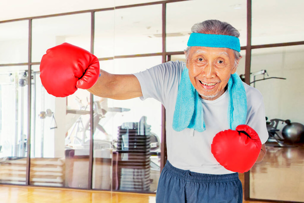Old man wearing boxing gloves while showing his punch and exercising in the fitness center - Foto, Bild