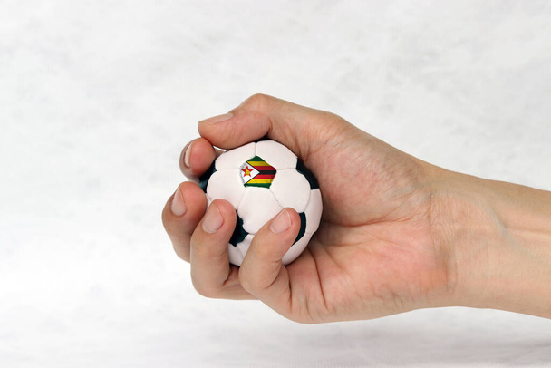 Mini fútbol en la mano y un punto negro del fútbol es la bandera de Zimbabue sobre fondo blanco. Concepto de deporte o el juego en el mango
. - Foto, imagen