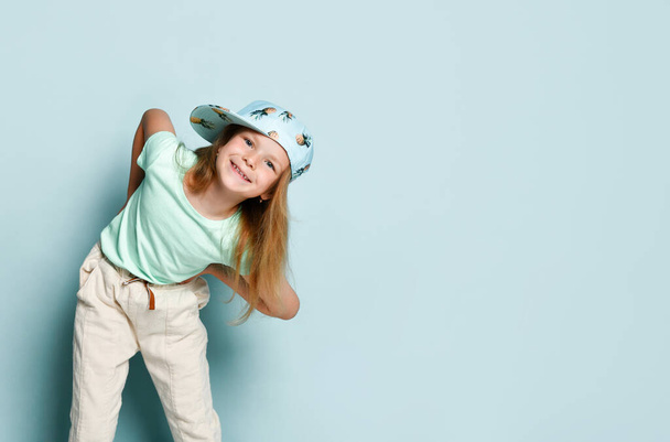 Kid in t-shirt, pants and cap printed by pineapples. She leaned forward with her arms wide apart, smiling, posing on turquoise background. Close up - Foto, immagini