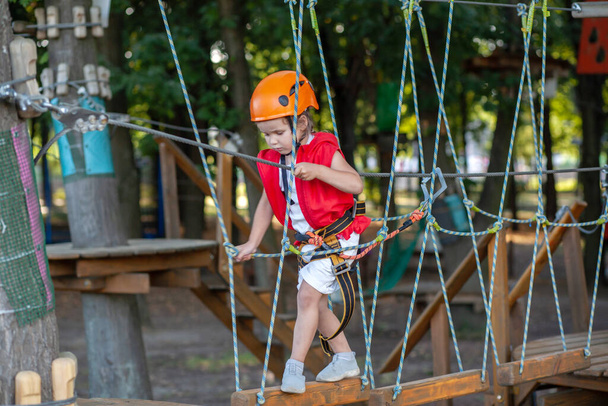 Uma menina em um capacete laranja e proteção especial passa por obstáculos em um parque de diversões corda, tempo de lazer das crianças, uma área de lazer para crianças, diversão no parque
. - Foto, Imagem