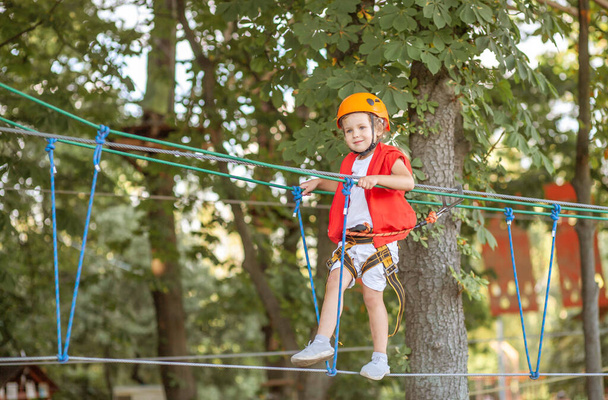 Ένα μικρό κορίτσι σε ένα πορτοκαλί κράνος και ειδική προστασία περνά από εμπόδια σε ένα πάρκο αναψυχής σχοινί, ελεύθερο χρόνο των παιδιών, ένα χώρο παιχνιδιού για νήπια, διασκέδαση στο πάρκο. - Φωτογραφία, εικόνα