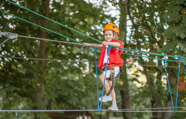Een klein meisje met een oranje helm en speciale bescherming gaat door obstakels in een touwattractiepark, kindervrije tijd, een speeltuin voor peuters, plezier in het park. - Foto, afbeelding