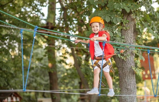 Маленькая девочка в оранжевом шлеме и специальной защите проходит через препятствия в веревочном парке развлечений, детского досуга, игровая площадка для малышей, развлечения в парке. - Фото, изображение