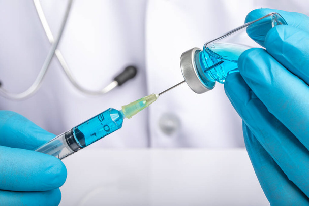 Un médecin en manteau blanc tirant un liquide bleu clair clair d'un flacon dans une seringue. Médicament ou administration du vaccin, concept de prévention covid-19.  - Photo, image