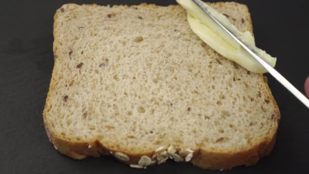 Butter is spread with a knife over a slice of cereal bread on a gray stone surface. Close-up - Footage, Video