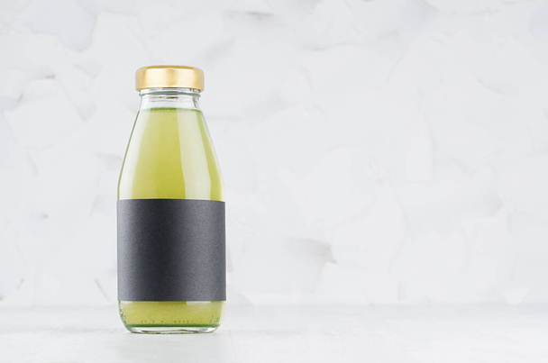 Green kiwi juice in glass bottle with gold cap and black blank label mock up on white wood table in light interior with copy space, template for packaging, advertising, design product, branding. - 写真・画像