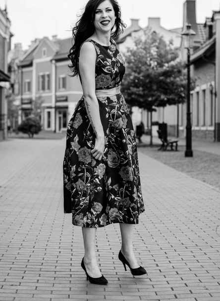 Gorgeous vintage brunette woman with curly hair on a style 50s in black dress with red roses. Pretty girl walk on a streets and have a good mood feeling wonderful - Foto, Bild