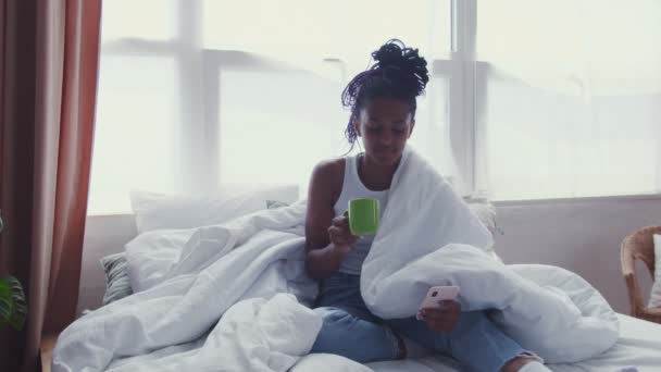 Vonzó fiatal afro-amerikai nő ül az ágyban, iszik reggeli kávét és használja a telefont - Felvétel, videó