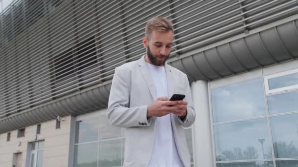 Een jonge zakenman leest een e-mail en verheugt zich over het succes van zijn bedrijf. Hij draagt een licht jasje en een wit T-shirt op de achtergrond van het kantoorcentrum. Succesvolle mensen, telefoon. - Video