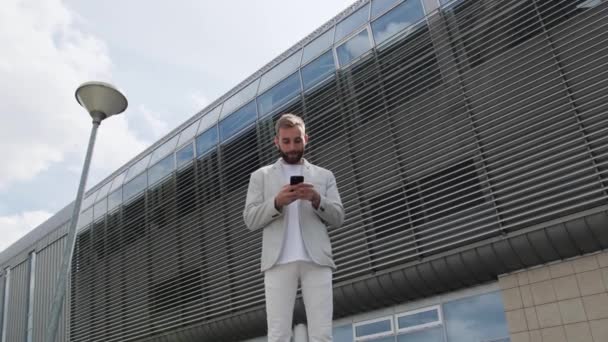 Молодой парень с бородой и в светлой куртке пользуется мобильным телефоном, улыбаясь на фоне современного здания. Одетые в светлую куртку и белую футболку. - Кадры, видео