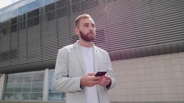 Jeune homme avec une barbe et dans une veste légère utilise une application mobile et réagit émotionnellement à l'extérieur. Jeux sur le téléphone, applications, navigateur. - Séquence, vidéo