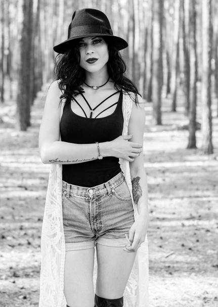 Gorgeous beautiful curly brunette woman with black hat and lace Bohemian cardigan, in boho style walk in forest and thinkin about life, near wood - Foto, afbeelding