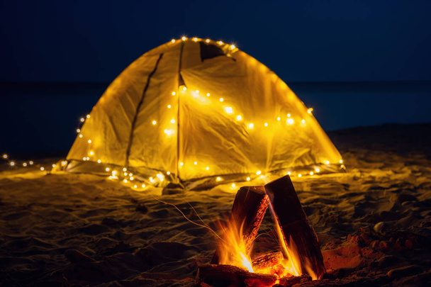 The fire at night on the beach. Summer mood. Night camping with a retro garland on sea shore. - Photo, Image
