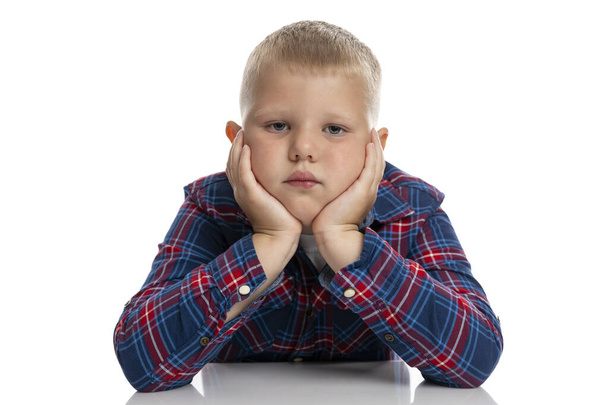 A fat, sad boy sits at a table with his head in his hands. Schoolboy in a plaid shirt. Back to school. Isolated on white background. Close-up. - Fotoğraf, Görsel
