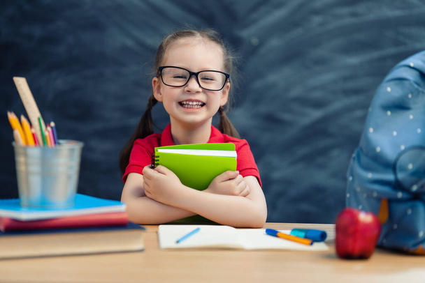 Back to school! Happy cute industrious child is sitting at a desk indoors. Kid is learning in class on background of blackboard. - Photo, image